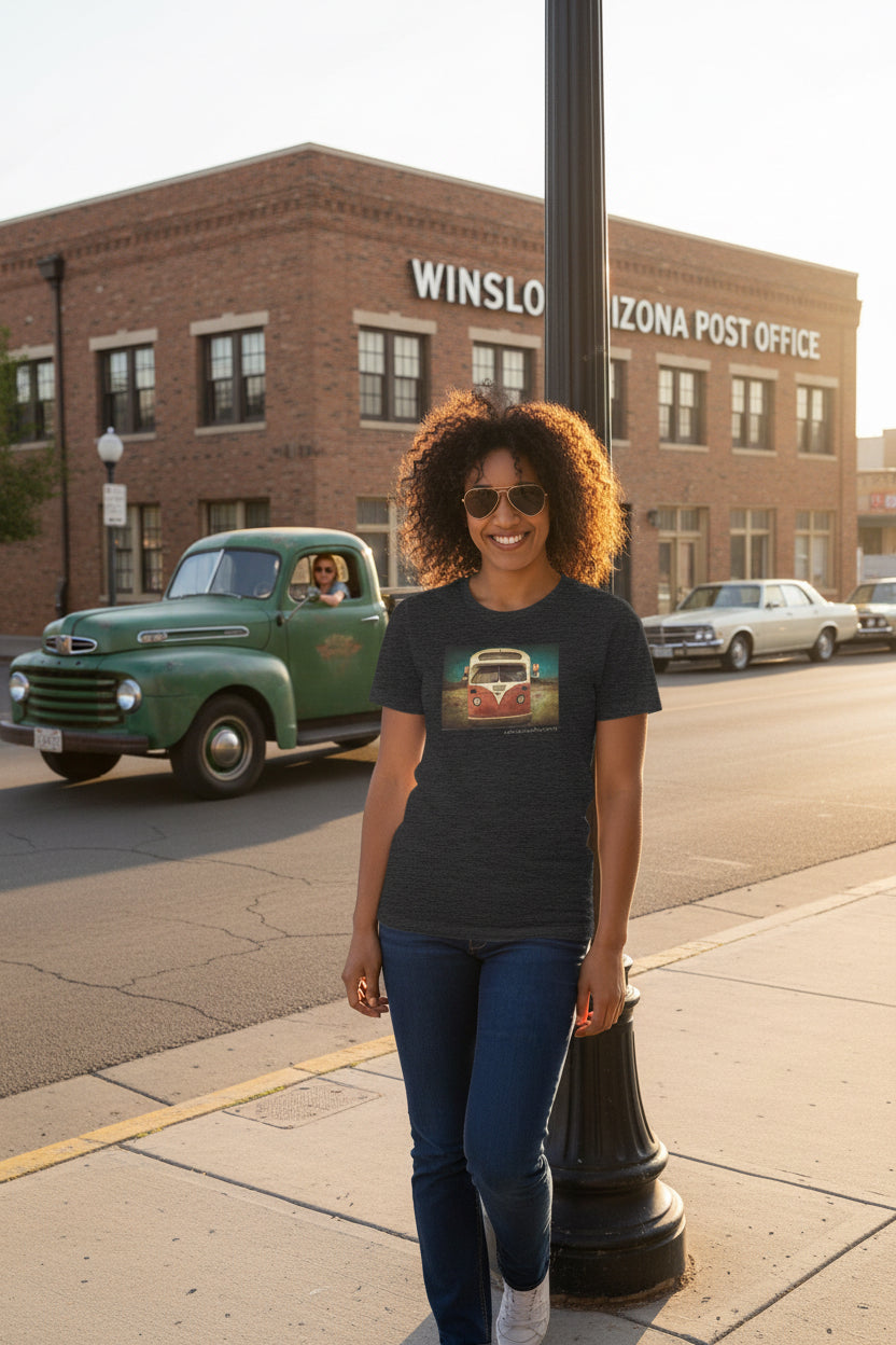 A woman wearing a dark heather gray graphic t-shirt with a retro school bus print, standing on a corner in front of a brick building labeled 'WINSLOW ARIZONA POST OFFICE'.