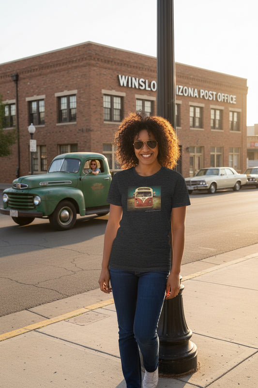 A woman wearing a dark heather gray graphic t-shirt with a retro school bus print, standing on a corner in front of a brick building labeled 'WINSLOW ARIZONA POST OFFICE'.